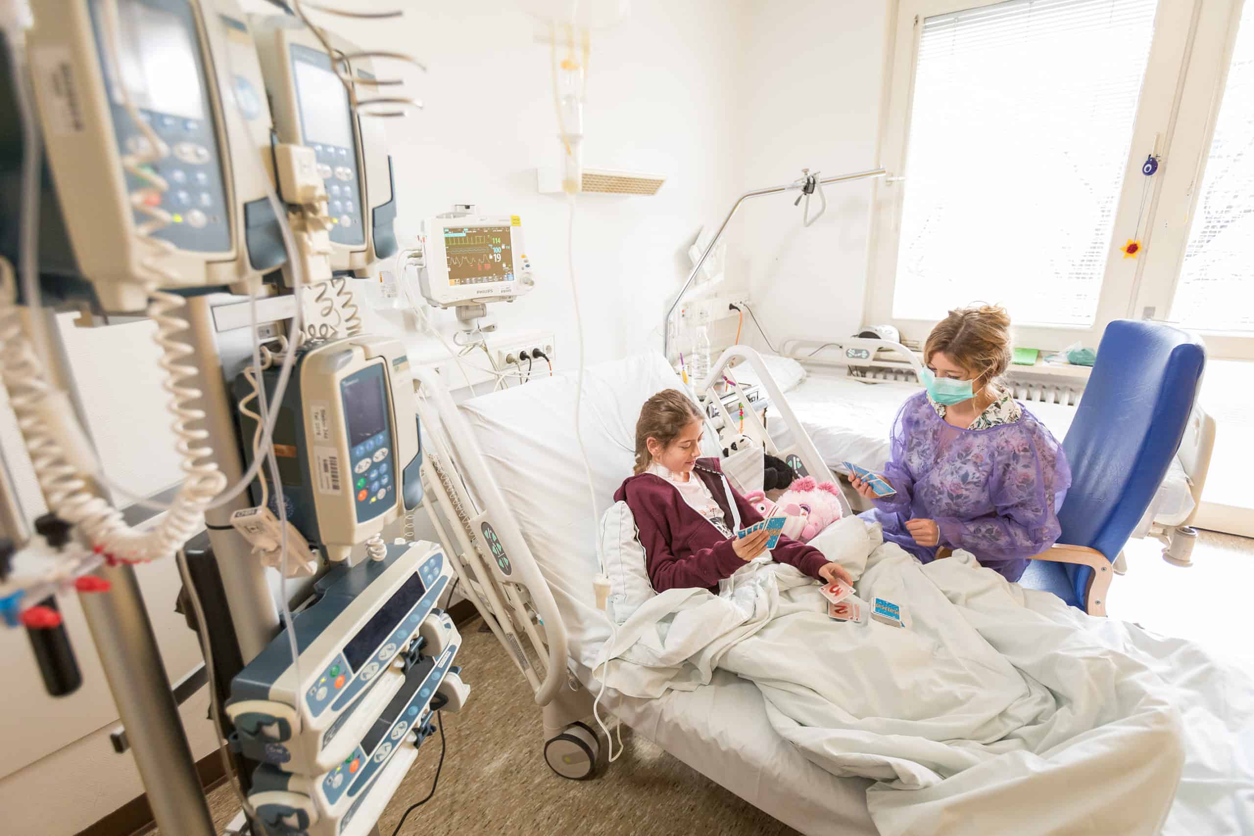 Kind im Bett neben vielen medizinischen Geräten. Daneben die Mutter mit Schutzmaske