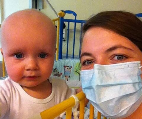 Baby im Kinderbett, daneben Mutter mit medizinischer Schutzmaske