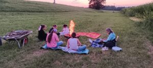 6 Personen sitzen im Abendlicht auf einer Wiese um eine Feuerschale