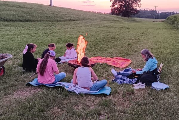 6 Personen sitzen im Abendlicht auf einer Wiese um eine Feuerschale
