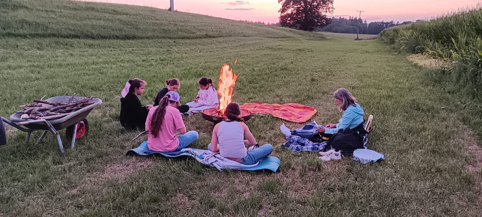 6 Personen sitzen im Abendlicht auf einer Wiese um eine Feuerschale