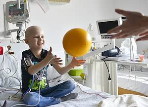 Kind sitzt in Klinikbett und fängt einen Ball angeschloßen an Schläuche, Kabel und medizinische Maschinen