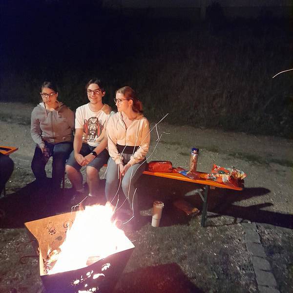 3 junge Leute sitzen nachts auf 1 Bierbank am Feuerkorb